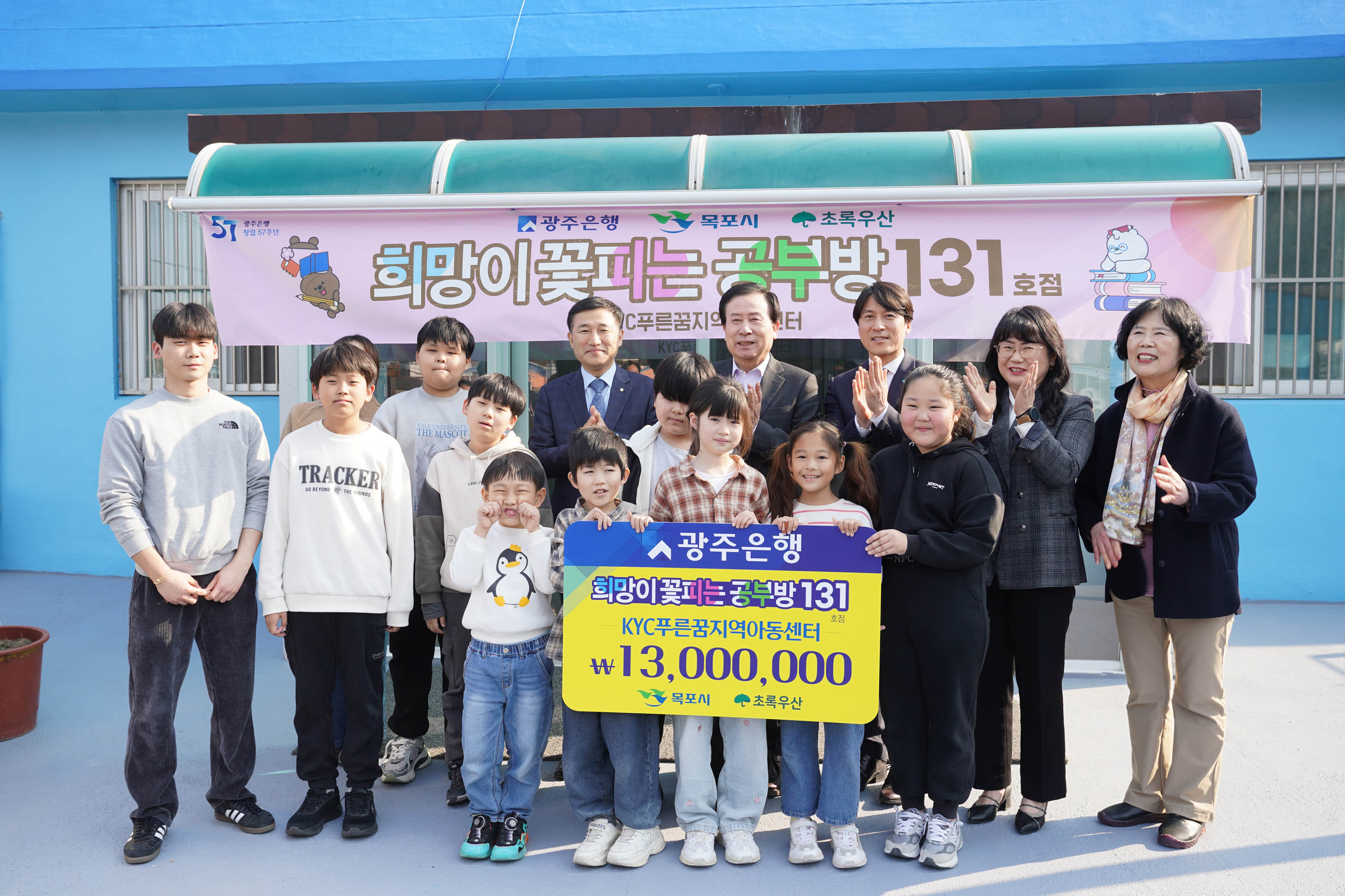 여러 명의 어린이들과 박홍률 목포시장과 4명의 관계자와 함께 서 있는 단체 사진. 어린이들이 '광주은행 희망이꽃피는공부방131 KYC 푸른꿈지역아동센터 13,000,000 목포시 초록우산' 문구가 적힌 팻말을 들고 있다. 배경에는 '광주은행 목포시 초록우산 희망이 꽃피는공부방 131호점 푸름꿈지역센터' 라는 현수막이 걸려있다.