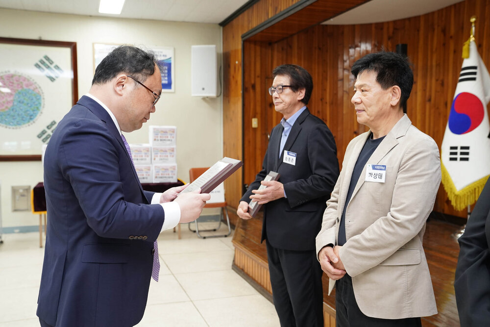 정장을 입고 안경을 낀 남성이 베이지색 자켓을 입은 남성에게 패를 수여하고 있다