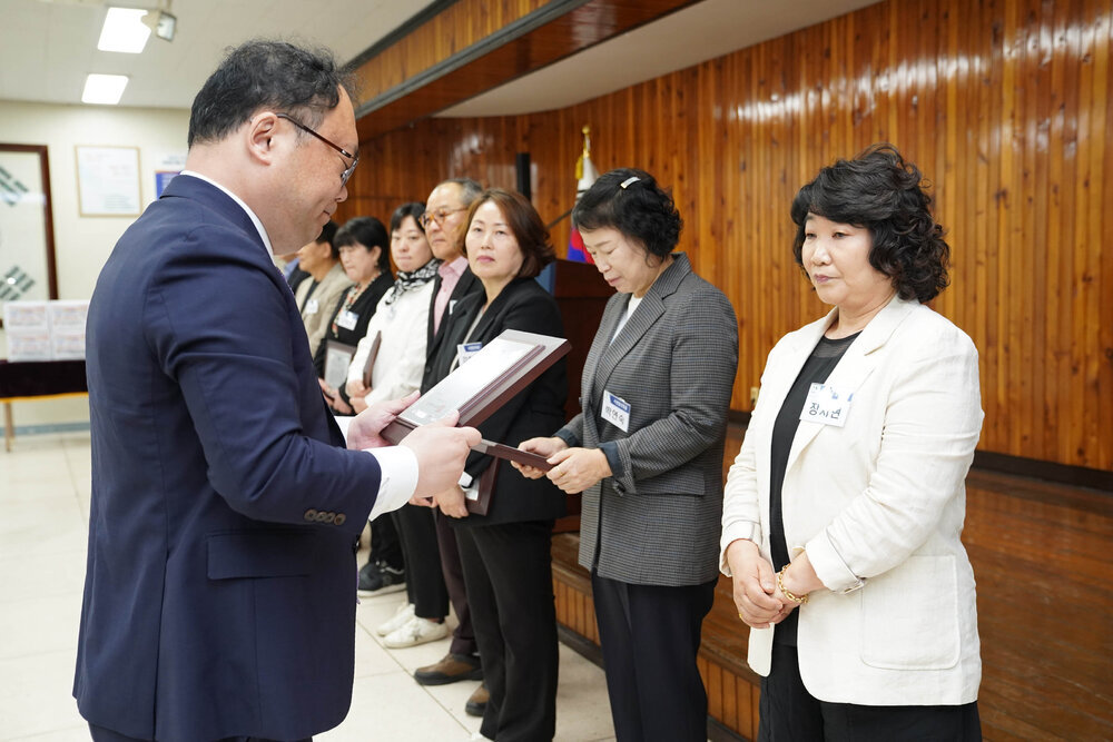 정장을 입고 안경을 낀 남성이 흰색 자켓을 입은 여성에게 패를 수여하고 있다
