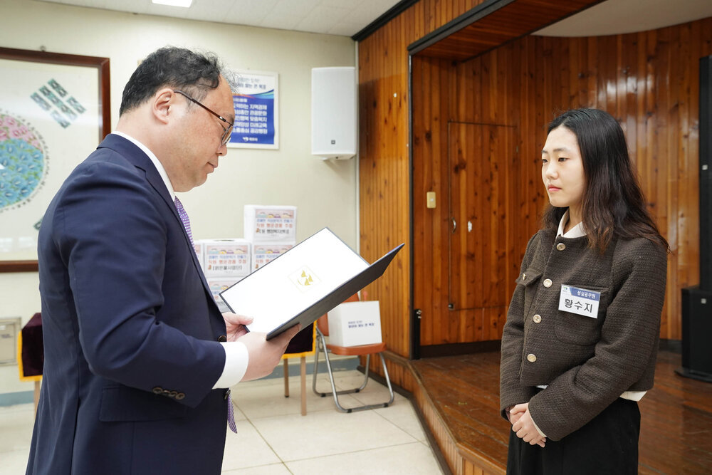정장을 입고 안경을 낀 남성이 갈색 트위드 자켓을 입은 황수지에게 상장을 수여하고 있다
