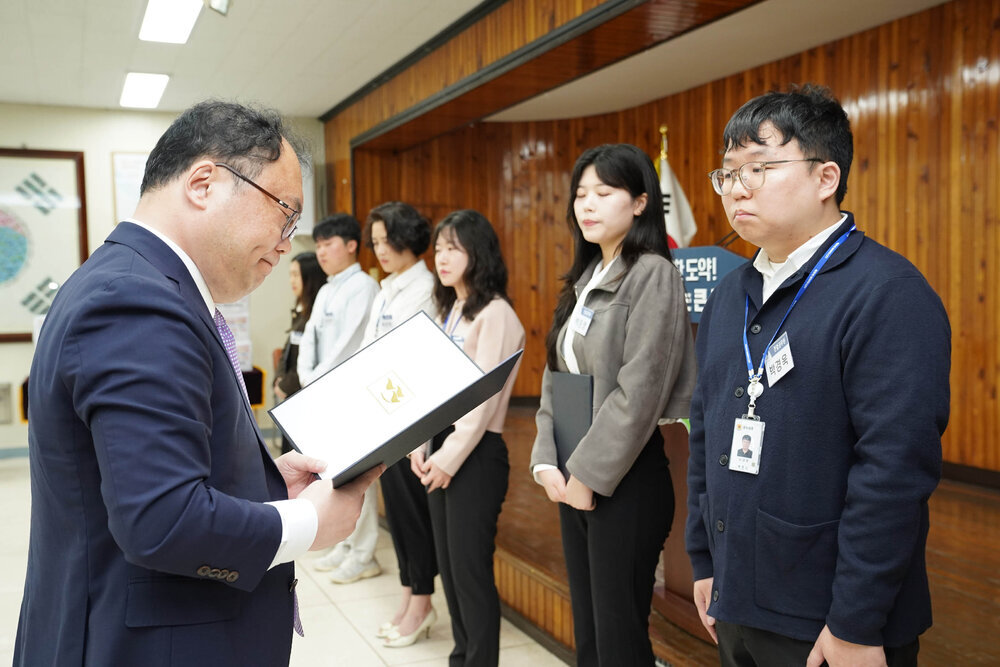 정장을 입고 안경을 낀 남성이 남색 자켓을 입은 남성에게 상장을 수여하고 있다