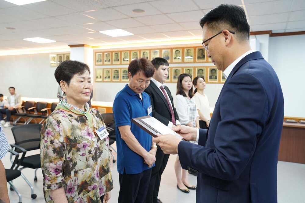 정장 차림의 관계자가 꽃무늬 상의를 입은 여성 수상자에게 표창패를 전달하며 읽고 있는 모습이다.