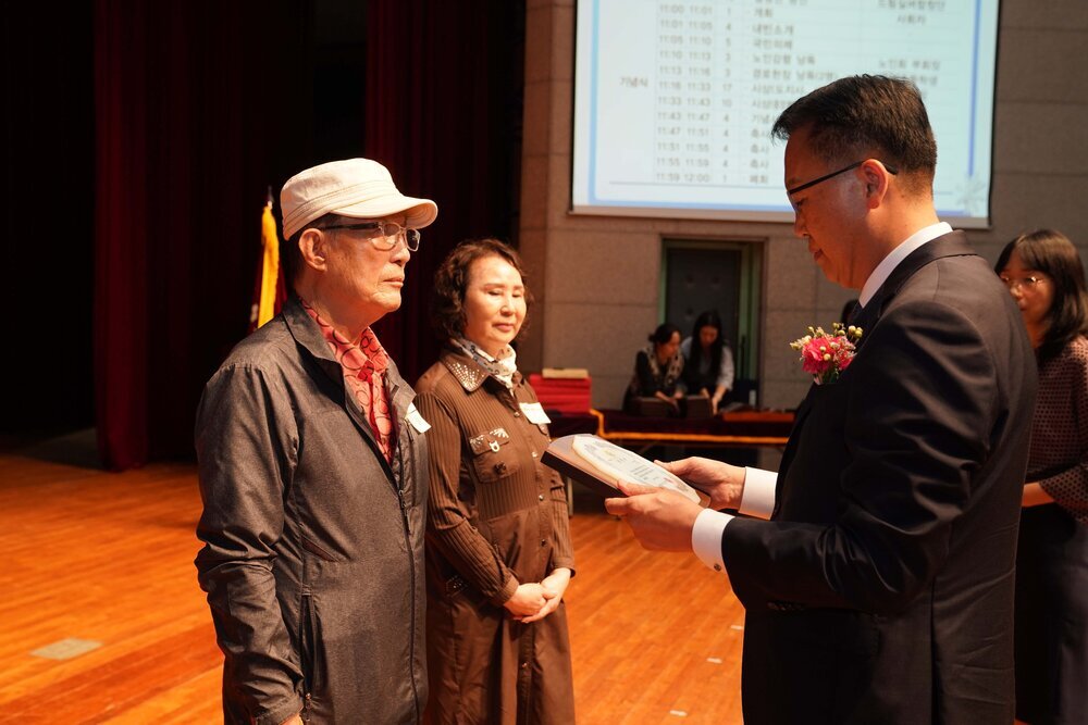 정장 차림의 관계자가 모자를 착용한 남성 수상자에게 패를 전달하며 읽고 있는 모습이다.