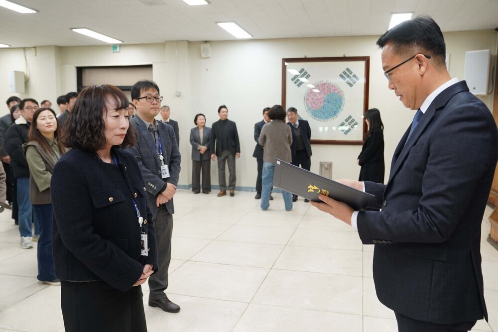 2026년 상반기 4급이하 정기인사로 정장을 입은 남성이 실내 행사장에서 한 여성에게 상장을 전달하며 내용을 읽고 있고, 여성은 파마머리에 검은색 정장 자켓을 입고 사원증을 착용한 채 두 손을 모은 상태로 서 있으며 뒤쪽으로 참석자들이 줄지어 서서 행사를 지켜보고 있다.