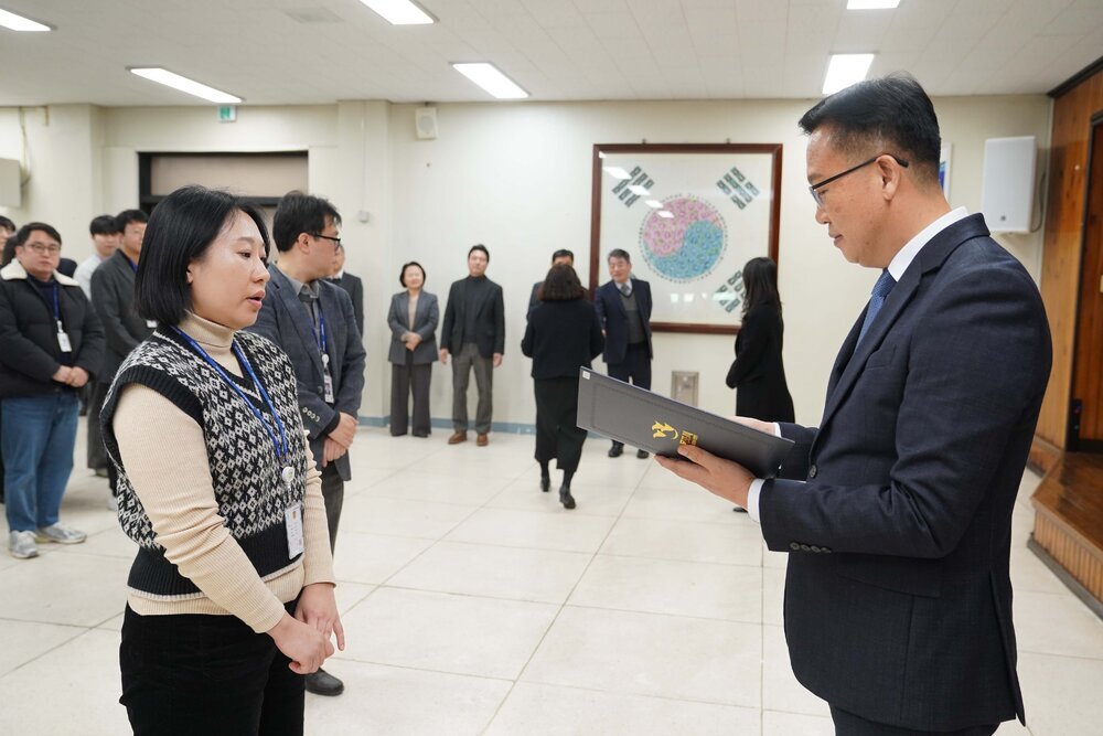 2026년 상반기 4급이하 정기인사로 정장을 입은 남성이 실내 행사장에서 한 여성에게 상장을 전달하며 내용을 읽고 있고, 여성은 베이지색 목폴라 위에 기하학 무늬 베스트를 입고 사원증을 착용한 채 두 손을 모은 상태로 서 있으며 뒤쪽으로 참석자들이 줄지어 서서 행사를 지켜보고 있다.