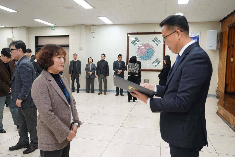 2026년 상반기 4급이하 정기인사로 정장을 입은 남성이 실내 행사장에서 한 여성에게 상장을 전달하며 내용을 읽고 있고, 여성은 갈색 파마머리에 갈색 자켓을 입고 사원증을 착용한 채 두 손을 모은 상태로 서 있으며 뒤쪽으로 참석자들이 줄지어 서서 행사를 지켜보고 있다.