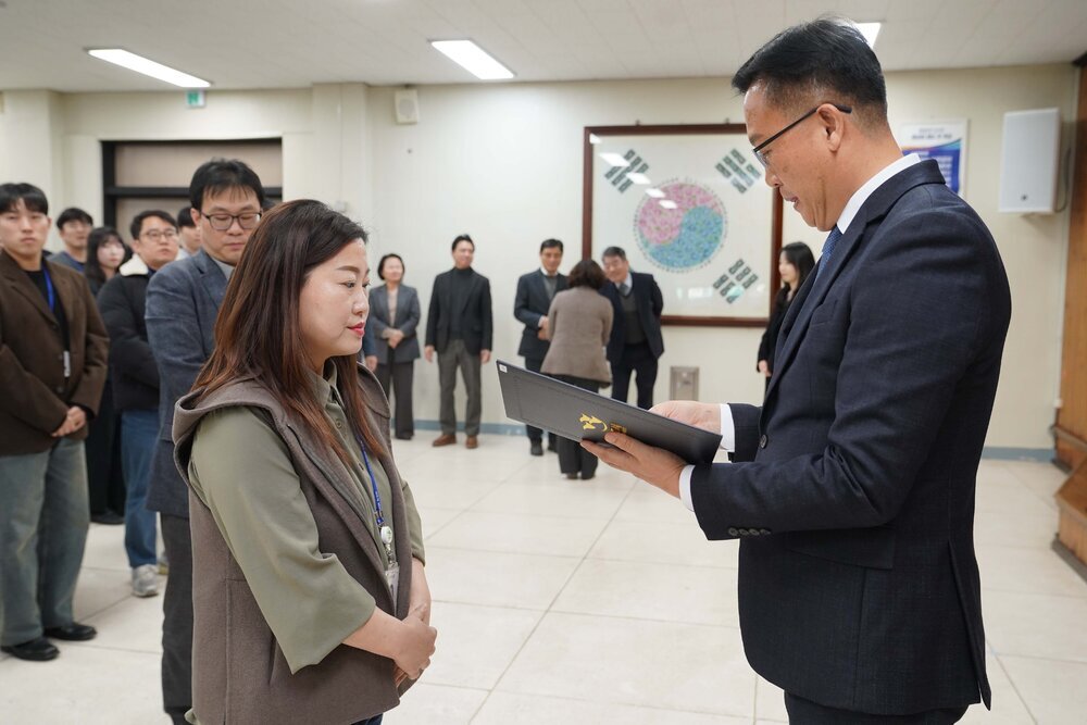 2026년 상반기 4급이하 정기인사로 정장을 입은 남성이 실내 행사장에서 한 여성에게 상장을 전달하며 내용을 읽고 있고, 여성은 긴 생머리에 카키색 조끼를 겹쳐 입고 사원증을 착용한 채 두 손을 모은 상태로 서 있으며 뒤쪽으로 참석자들이 줄지어 서서 행사를 지켜보고 있다.