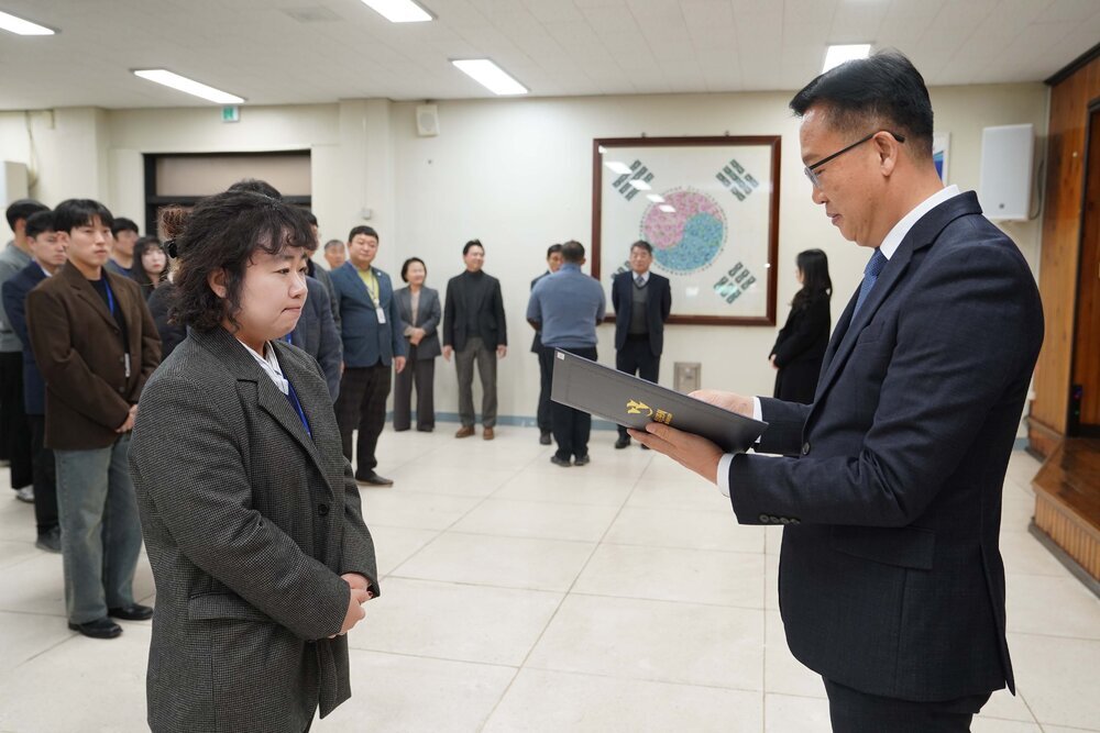 2026년 상반기 4급이하 정기인사로 정장을 입은 남성이 실내 행사장에서 한 여성에게 상장을 전달하며 내용을 읽고 있고, 여성은 묶은 파마머리에 짙은 회색 자켓을 입고 두 손을 모은 상태로 서 있으며 뒤쪽으로 참석자들이 줄지어 서서 행사를 지켜보고 있다.