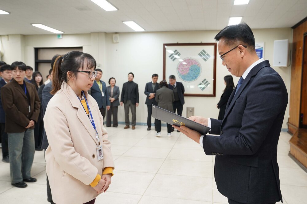 2026년 상반기 4급이하 정기인사로 정장을 입은 남성이 실내 행사장에서 한 여성에게 상장을 전달하며 내용을 읽고 있고, 여성은 안경을 쓰고 머리를 묶은 채 밝은 베이지색 자켓을 입고 사원증을 착용한 채 두 손을 모은 상태로 서 있으며 뒤쪽으로 참석자들이 줄지어 서서 행사를 지켜보고 있다.