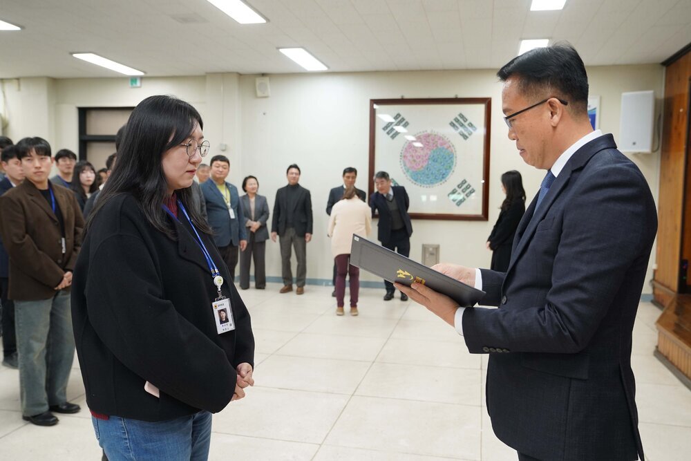2026년 상반기 4급이하 정기인사로 정장을 입은 남성이 실내 행사장에서 한 여성에게 상장을 전달하며 내용을 읽고 있고, 여성은 안경을 쓰고 검은색 코트와 청바지를 입고 사원증을 착용한 채 두 손을 모은 상태로 서 있으며 뒤쪽으로 참석자들이 줄지어 서서 행사를 지켜보고 있다.