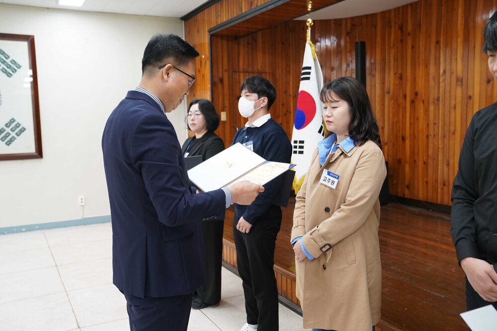 4월 정례조회에 정장을 입고 안경을 낀 남성이 베이지색 트렌치코트를 입은 고주원에게 패를 수여하고 있다
