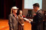 정장 차림의 관계자가 모자를 착용한 남성 수상자에게 패를 전달하며 읽고 있는 모습이다.