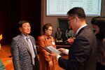 정장 차림의 관계자가 회색 정장을 입은 남성 수상자에게 패를 전달하며 읽고 있는 모습이다.