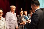 정장 차림의 관계자가 회색 재킷을 입은 남성 수상자에게 패를 전달하며 읽고 있는 모습이다.