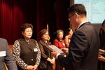 정장 차림의 관계자가 검은색 조끼를 입은 여성 수상자에게 패를 전달하며 읽고 있는 모습이다.