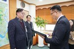 2026년 상반기 4급이하 정기인사로 정장을 입은 남성이 실내 사무실에서 한 남성에게 상장을 전달하며 내용을 읽고 있고, 남성은 안경을 쓰고 남색 자켓에 와인색 니트를 겹쳐 입은 채 사원증을 착용하고 서 있으며 왼쪽 벽면에는 대형 지도가 걸려 있다.
