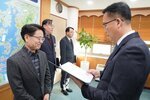 2026년 상반기 4급이하 정기인사로 정장을 입은 남성이 실내 사무실에서 한 남성에게 상장을 전달하며 내용을 읽고 있고, 남성은 안경을 쓰고 회색 체크무늬 자켓을 입은 채 두 손을 모으고 서 있으며 뒤쪽으로 다른 참석자들이 지켜보고 있다.