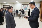 2026년 상반기 4급이하 정기인사로 정장을 입은 남성이 실내 행사장에서 한 남성에게 상장을 전달하며 내용을 읽고 있고, 남성은 안경을 쓰고 짙은 회색 보풀 니트 자켓을 입은 채 두 손을 내린 상태로 서 있으며 뒤쪽으로 참석자들이 줄지어 서서 행사를 지켜보고 있다.